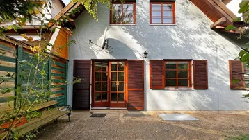 Rückseite des Hauses mit gepflasterter Terrasse, Holztüren und Fensterläden, umgeben von Bäumen.
