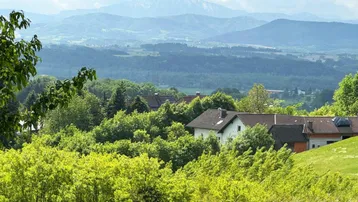 Landschaftliche Ansicht mit Häusern, umgeben von viel Grün und Bergen im Hintergrund.