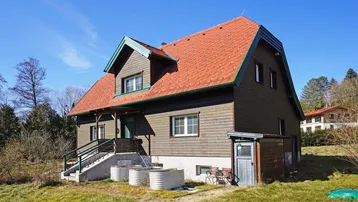 Ländliches Einfamilienhaus mit Holzfassade, rotem Dach und großem Gartenbereich unter blauem Himmel.