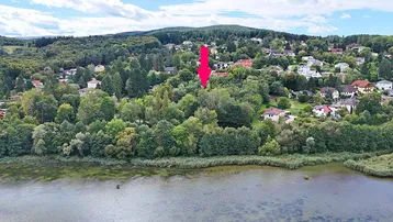Luftaufnahme eines Sees mit bewaldeten Ufern und einer Wohnsiedlung im Hintergrund, markiert mit einem roten Pfeil.