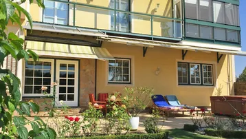 Gelbe Hausfassade mit möblierter Terrasse, Markise und gepflegtem Gartenbereich.