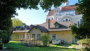 Blick auf das charmante Gebäude mit Garten und Terrasse, umgeben von üppigem Grün und einer Kirche im Hintergrund.