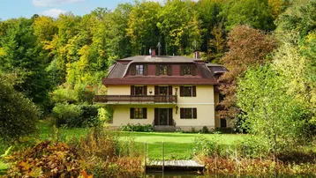Traditionelles Haus mit Mansarddach und Balkon, umgeben von üppiger Natur und einem Teich.