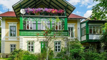 Historisches Gebäude mit auffälliger grüner Holzveranda und blühenden Blumenkästen im Vorgarten.