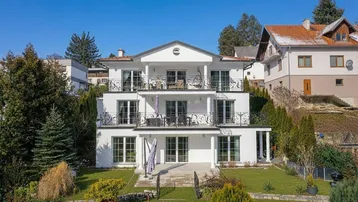Elegantes weißes Mehrfamilienhaus mit mehreren Balkonen, Terrassen und gepflegtem Gartenbereich unter blauem Himmel.