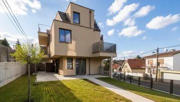Modernes Mehrfamilienhaus mit beiger Fassade, Balkonen und gepflegtem Vorgarten unter blauem Himmel.