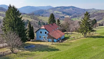 Blaues Haus mit rotem Dach in ländlicher Umgebung, umgeben von grünen Hügeln und Bäumen.