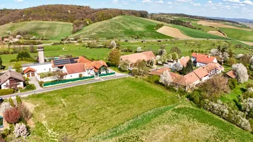 Luftaufnahme einer ländlichen Siedlung mit grünen Feldern und bewaldeten Hügeln unter blauem Himmel.