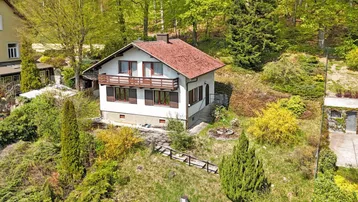 Freistehendes Einfamilienhaus mit rotem Ziegeldach und Balkon, umgeben von viel Grün und Bäumen.