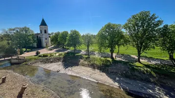 Panoramablick auf einen Flusslauf mit üppigem Grün und einer Kirche im Hintergrund unter blauem Himmel.