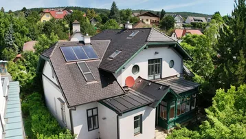 Hausansicht von oben mit Satteldach, Solarpaneelen und umgeben von viel Grün.