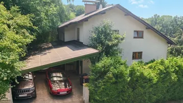 Hausfassade mit integriertem Carport für zwei Fahrzeuge, umgeben von üppigem Grün.