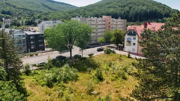 Panoramablick auf eine Straße mit Gebäuden, einem unbebauten Grundstück und bewaldeten Hügeln im Hintergrund.