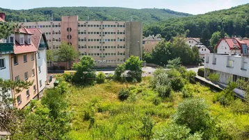Blick auf ein unbebautes Grundstück mit umliegenden Wohngebäuden und bewaldeten Hügeln im Hintergrund.