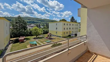 Blick vom Balkon auf die umliegenden Gebäude, einen Spielplatz und grüne Hügel in der Ferne.