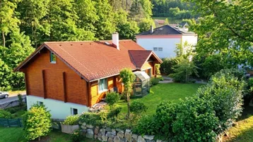 Charmantes Holzhaus mit rotem Ziegeldach und gepflegtem Garten in grüner, waldreicher Umgebung.