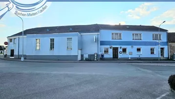 Gebäudeansicht eines blauen Hauses mit großem Vorplatz und Werbebanner der Immobilienfirma.