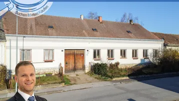 Langes, weißes Gebäude mit braunem Ziegeldach und mehreren Fenstern, umgeben von spärlicher Vegetation.