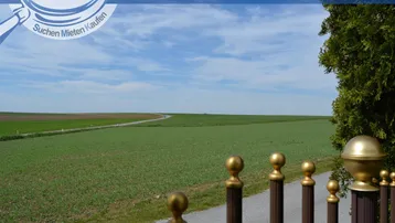 Weitläufiger Blick über grüne Felder und die ländliche Umgebung, begrenzt durch einen Zaun im Vordergrund.