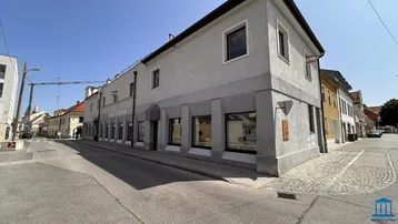 Graues Gebäude an einer Straßenecke mit großen Schaufenstern und einer Kopfsteinpflasterstraße unter blauem Himmel.