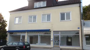 Gebäudeansicht mit Ladenflächen im Erdgeschoss und Wohnungen darüber, geprägt von einer hellen Fassade und blauen Markisen.