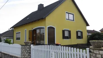 Gelbes Einfamilienhaus mit Satteldach, weißem Zaun und braunen Fensterläden.