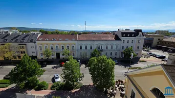 Panoramablick über eine Stadtstraße mit historischen Gebäuden und grünen Bäumen unter blauem Himmel.