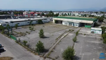 Luftaufnahme eines weitläufigen Gewerbegrundstücks mit mehreren Gebäuden, großen Freiflächen und Bergblick.
