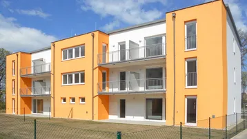 Modernes Mehrfamilienhaus mit leuchtend orangefarbener Fassade, großen Fenstern und Balkonen unter blauem Himmel.