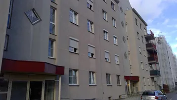 Graue Fassade eines Mehrfamilienhauses mit vielen Fenstern, Rollläden und Balkonen unter bewölktem Himmel.