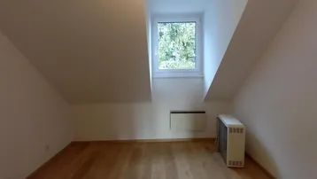 Heller Dachbodenraum mit schrägen Wänden, Holzfußboden und einem Fenster mit Blick ins Grüne.