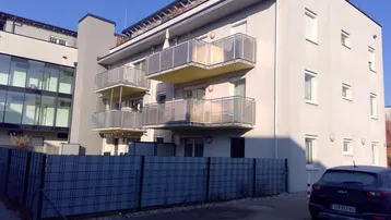 Modernes Mehrfamilienhaus mit hellgrauer Fassade und mehreren Balkonen mit Glasgeländer.