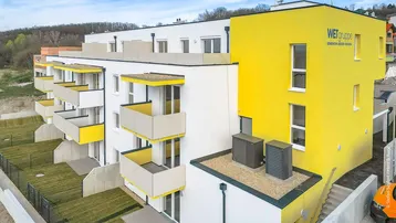 Modernes Mehrfamilienhaus mit weißer und gelber Fassade sowie mehreren Balkonen und Grünflächen.