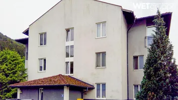 Mehrfamilienhaus mit heller Fassade, Satteldach und kleinen Balkonen, umgeben von Bäumen.