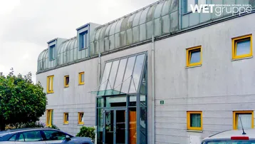 Modernes Gebäude mit markanter Glasfassade und gelben Fensterrahmen unter bewölktem Himmel.