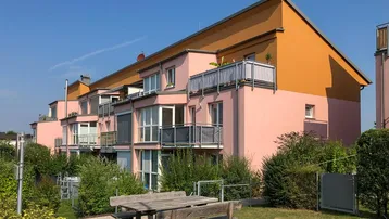 Mehrfamilienhaus mit rosa Fassade, Balkonen und einem gemeinschaftlichen Gartenbereich mit Sitzgelegenheiten.