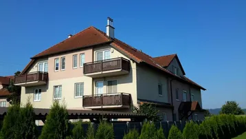 Mehrfamilienhaus mit mehreren Balkonen und Satteldach, umgeben von grüner Landschaft.