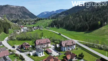 Luftaufnahme eines Wohngebäudes in einer grünen Berglandschaft mit Straßen und umliegenden Häusern.