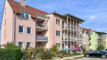 Mehrfamilienhaus mit rosa Fassade, Balkonen und gepflegter Außenanlage unter blauem Himmel.