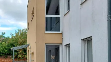 Moderne Gebäudefassade mit hellen Putzflächen, großen Fenstern und einem barrierefreien Zugang über eine Rampe.