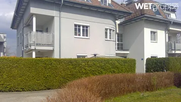 Graue Fassade eines Mehrfamilienhauses mit Balkonen und gepflegter Hecke im Vordergrund.