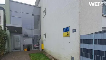 Moderner Eingangsbereich eines Gebäudes mit Glasfassade, weißem Putz und gepflegter Umgebung.