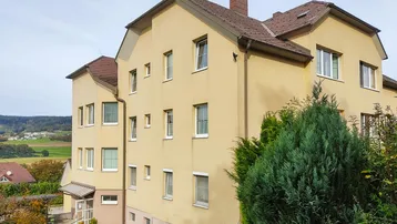 Gelbes Mehrfamilienhaus mit Satteldach und grüner Umgebung unter einem klaren Himmel.