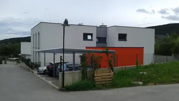 Moderne Wohnanlage mit weißer und oranger Fassade, Flachdach und Carport in ländlicher Umgebung.