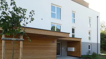 Moderne Wohnhausfassade mit weißem Putz und Holzverkleidung am Eingangsbereich, umgeben von Grün.
