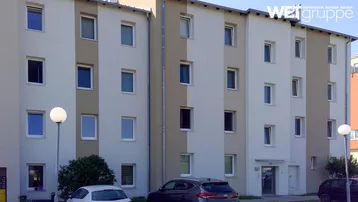 Mehrstöckiges Wohngebäude mit heller Fassade und zahlreichen Fenstern, umgeben von Parkplätzen und Grünflächen.