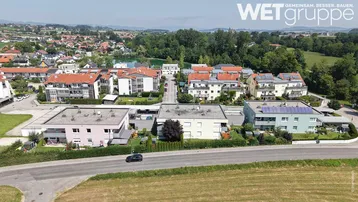 Luftaufnahme einer gepflegten Wohnsiedlung mit modernen Gebäuden, Photovoltaikanlagen und Blick auf einen See sowie die umliegende grüne Landschaft.