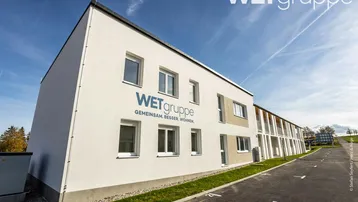 Modernes Mehrfamilienhaus mit weißer Fassade und Parkplätzen im Freien unter blauem Himmel.