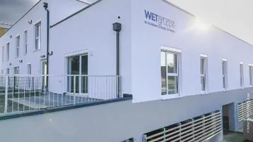 Modernes Gebäude mit weißer Fassade, großen Fenstern und einem Balkon im ersten Stock.