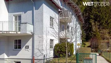 Mehrfamilienhaus mit heller Putzfassade und mehreren Balkonen, umgeben von Bäumen und Grünflächen in ländlicher Umgebung.